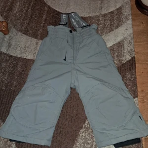 Grå täckbyxor med hängslen - Grå täckbyxor från SIS 75 med justerbara hängslen och förstärkta knän. Byxorna har dragkedja och kardborre i midjan, samt reflexdetalj på benet för extra synlighet. Fodrade och perfekta för kalla dagar. Snygg och funktionell design.