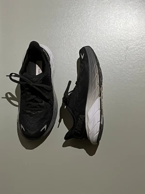 Hoka Arahi 6 - Säljer ett par svarta Hoka sneakers med vit och grå tjock sula. Skorna har snörning och rund tå, med Hoka-logga på sidan och bak. Ovandelen är i syntetmaterial och mesh för bra ventilation. Perfekta för dig som gillar att träna eller vill ha riktigt sköna skor. Endast använt vid fåtal tillfällen och är i sparsamt använt skick!