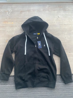 Svart hoodie från Polo Ralph Lauren - Svart hoodie från Polo Ralph Lauren med vit broderad logga på bröstet och vita snören i huvan. Tröjan har dragkedja framtill, ribbade muddar och är tillverkade bra material. Klassisk och clean look med normal passform och lång ärm.