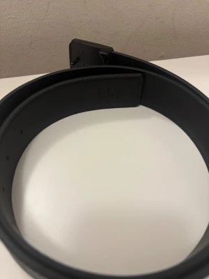 Svart Louis Vuitton skärp i skinn - Säljer ett stilrent svart Louis Vuitton bälte i skinn med präglade LV-loggor och monogram längs hela remmen. Bältet har en matt svart LV-spänne som ger en lyxig och modern vibe. Perfekt accessoar för att lyfta din outfit.