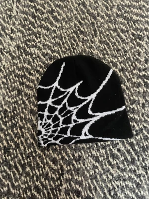 Y2k Svart mössa med spindelnätsmönster - Cool svart mössa med vitt spindelnätsmönster på ena sidan. Perfekt accessoar för dig som gillar edgy och unika detaljer. Mössan är mjuk och stretchig, passar de flesta huvudstorlekar. Grym till höst- och vinteroutfits.