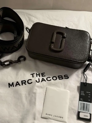 Svart Väska  - Säljer en svart axelväska från Marc Jacobs med avtagbar bred axelrem och ikonisk logga framtill. Perfekt storlek för det viktigaste och stängs med dragkedja. Märkesdetaljer på både rem och väska. Kartong, äkthetsbevis/kort, dustbag medföljer. Pris kan diskuteras. 