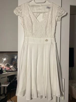 Vit klänning från Chiara Forthi - Vit studentklänning från Chiara Forthi med spets upptill, v-ringning och korta ärmar. Kjolen är lätt utsvängd och materialet känns mjukt och följsamt. Baksidan har öppen rygg med knappar i nacken.
