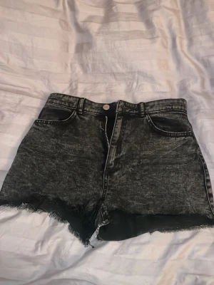 Svarta jeansshorts - Säljer ett par svarta jeansshorts med råa, fransiga kanter och slitna detaljer. Shortsen har klassisk femficksmodell, bälteshällor och dragkedja framtill. Dom har aldrig kommit till användning 