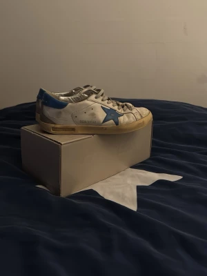 Golden Goose vita sneakers med blå detalj - Golden Goose sneakers i vitt skinn med blå stjärna på sidan och blå hälkappa. Grå snörning och beige sula ger en cool vintage vibe. Klassisk låg modell med rund tå och platt sula. Perfekta för dig som gillar streetstyle och unika detaljer. Vid frågor är det bara att kontakta! 😊