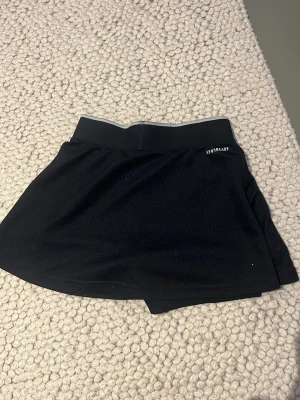 Svarta Adidas skorts med reflexdetalj - Säljer en tjol från Adidas med bred midjeresår och reflexdetalj upptill. Materialet är lätt och andas, perfekt för träning eller tennis. Diskret Adidas-logga framtill och AEROREADY-tryck på sidan. Skorts-modell med inbyggda shorts under kjolen.