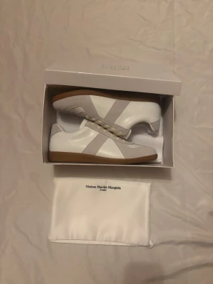 Maison Margiela Skor - Maison Margiela sneakers i vitt skinn och ljusgrå mocka med klassisk gummisula. Snygg retrostil med rund tå och krämvita snören. Kommer med originalkartong och dustbag. Perfekta för dig som gillar minimalistisk och clean design.