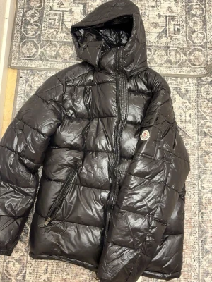 Svart Moncler pufferjacka med huva - Säljer en svart pufferjacka från Moncler med glansig finish och stor huva. Jackan har quiltad design, dragkedja framtill och två snedställda fickor med dragkedja. Klassisk Moncler-logga på ärmen. Perfekt för kalla dagar.