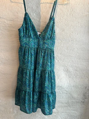 Turkos mönstrad sommarklänning - Säljer en kortklänning med paisleymönster i blått och turkost. Klänningen har smala axelband, v-ringning och volangdetaljer i kjolen. Perfekt för varma dagar och har en luftig passform.