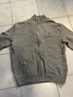 Grå kofta från Polo Ralph Lauren - Säljer en stilren grå kofta från Polo Ralph Lauren i mjuk pima bomull. Tröjan har dragkedja framtill, två fickor och den klassiska broderade loggan på bröstet. Perfekt till lager på lager och funkar året runt.