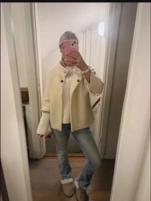 Beige kappa med knappar - Säljer en stilren beige kappa i ullblandning med hög krage och stora svarta knappar framtill. Jackan har en rak och något boxig passform med trekvartärmar. Perfekt för dig som gillar en clean och klassisk look.