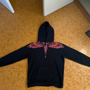 Svart hoodie med röda vingar från Marcelo Burlon - Hej jag säljer nu en exklusiv Marcelo Burlon “Wings” (Pris kan diskuteras vid snabba affärer) hoodie i svart med ikoniskt vingtryck i rött. Passar både vardag och fest. Väldigt fint skick. Storlek: [M/L]. Nypris ca 2500 - 3000 kr. Öppen för byten i storlek S/M. Pris kan diskuteras vid snabba affärer  Hör av dig vid intresse eller om ni har några flera funderingar! 