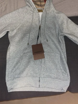 Grå hoodie från Burberry med dragkedja - Snygg grå hoodie från Burberry med klassiskt rutigt foder i huvan och dragkedja framtill. Tröjan har snörning i huvan, fickor på sidorna och diskret Burberry-logga broderad på bröstet. Perfekt för dig som vill ha en stilren och exklusiv look och den är helt ny som ni ser