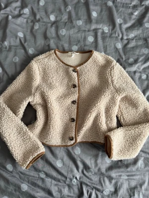 Beige teddyjacka med knappar - Mysig beige teddyjacka med bruna detaljer och knappar framtill. Jackan är croppad och har långärmad design. Perfekt för dig som vill ha en varm och trendig look under höst och vinter.