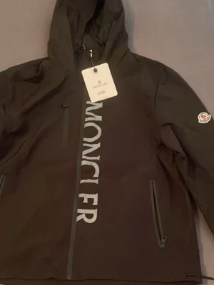 Moncler windbreaker  - Säljer en svart vindjacka från Moncler i storlek XL. Jackan har huva, stora Moncler-trycket längs dragkedjan, två sidofickor med dragkedja och en bröstficka. Tillverkad i slitstarkt material och har klassisk Moncler-logga på ärmen.