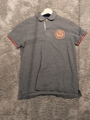Grå pikétröja från Tommy Hilfiger - Säljer en grå pikétröja från Tommy Hilfiger med klassisk krage och korta ärmar. Tröjan har en snygg broderad patch på bröstet och röda samt vita detaljer på ärmsluten. Perfekt till en avslappnad men ändå stilren look.