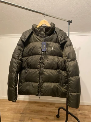 Mörkgrön pufferjacka från Polo Ralph Lauren - Snygg mörkgrön pufferjacka från Polo Ralph Lauren med broderad logga på bröstet. Jackan har hög krage, huva och dragkedja framtill samt dragkedjeförsedda fickor. Tillverkad i polyester och perfekt för kalla dagar.