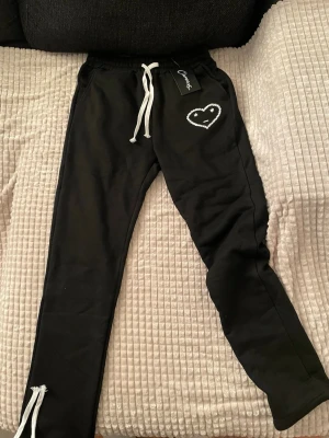 Svarta mjukisbyxor Carsicko hjärtdetalj - Svarta mjukisbyxor från Carsicko med vit snörning i midjan och ett broderat hjärta med ansikte på vänster sida. Byxorna har elastisk midja, ficka bak och är gjorda i mjukt material. Perfekta för chill dagar eller när du vill vara bekväm.