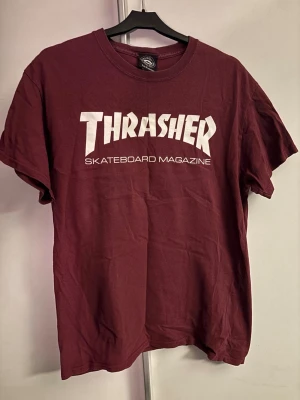 Vinröd Thrasher t-shirt - Säljer en vinröd Thrasher t-shirt med klassiskt vitt tryck på bröstet. Skön passform och rund hals. Perfekt för dig som gillar streetwear och skatekultur. Materialet är mjuk bomull och modellen är kortärmad.