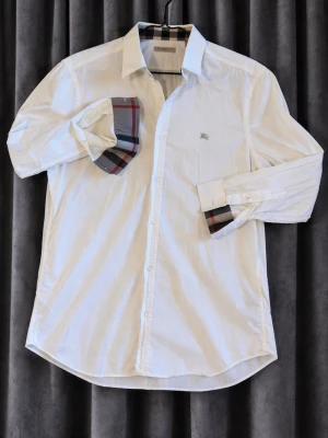 Burberry skjorta  - Klassisk vit skjorta från Burberry med ikoniskt rutigt tyg på insidan av manschetterna och kragen. Skjortan har knappar framtill, lång ärm och liten broderad logga på bröstet. Perfekt för dig som gillar stilrena detaljer och snygg design.