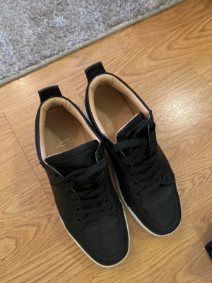 Svarta sneakers från Louboutin - Box ingår Snygga svarta sneakers från Louboutin i skinn med klassisk röd sula och vita kanter. Skorna har rund tå, svart snörning och diskret logga på innersulan. Insidan är beige och materialet känns lyxigt och mjukt.
