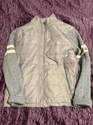 Grå moncler cardigan - Säljer en grå pufferjacka från Moncler med stickade, kabelmönstrade ärmar och vita ränder. Jackan har dragkedja framtill, Inga lappar på insidan av den, höga muddar och två fickor med dragkedja. Klassisk Moncler-logga på ena ärmen och riktigt skön passform. Passar mig bra som är 170