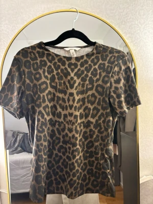Leopardmönstrad t-shirt från H&M - Säljer en snygg figurformad t-shirt från H&M med leopardmönster i bruna och svarta toner. T-shirten har korta ärmar och rund halsringning. Perfekt för dig som vill sticka ut lite extra med ett djurmönster i garderoben.