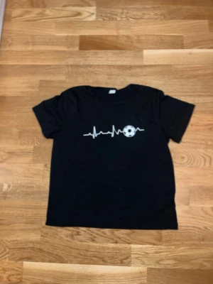 Svart t-shirt med fotbollstryck SHEIN - Svart t-shirt från SHEIN med korta ärmar och ett vitt tryck av en hjärtfrekvenslinje och en fotboll på bröstet. Tillverkad i mjuk bomull, perfekt för dig som gillar sportig stil.