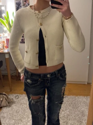 Krämvit kofta från Zara med guldknappar - Säljer en krämvit kofta från Zara med snygga guldknappar framtill. Koftan är stickad och har en croppad passform som ger en trendig look. Perfekt att slänga över en topp för en chic vibe.
