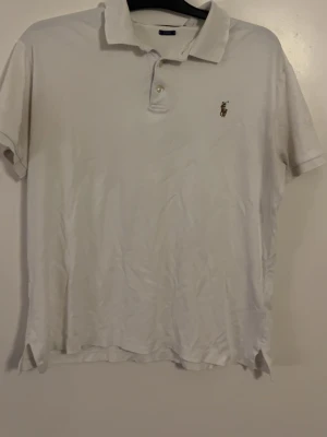Vit pikétröja från Ralph Lauren - Vit pikétröja från Ralph Lauren med klassisk krage och två knappar framtill. Tröjan har korta ärmar och den ikoniska broderade loggan på bröstet. Perfekt för en clean och stilren look.