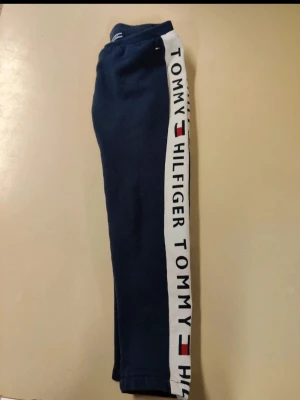 Blå mjukisbyxor Tommy Hilfiger - Mörkblå mjukisbyxor från Tommy Hilfiger med vit sidopanel där det står TOMMY HILFIGER och logga längs hela benet. Byxorna har en avslappnad passform och är tillverkade i mjukt bomullsmaterial, perfekta för chill dagar.