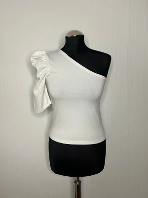 Vit oneshoulder topp från Monki - Säljer en vit oneshoulder topp från Monki i mjuk bomull. Toppen har en puffig, kort ärm på ena sidan och är ärmlös på andra. Snygg asymmetrisk design som ger en cool och trendig look. Perfekt att styla med jeans eller kjol.