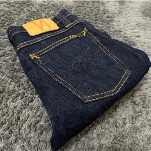 Mörkblå Nudie Jeans  - Säljer ett par mörkblå jeans från Nudie Jeans med klassisk gul kontrastsöm och slim fit. Jeansen har fem fickor och en snygg läderpatch bak i midjan. Tillverkade i slitstarkt jeansmaterial som sitter bra och ger en clean look