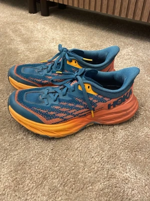 Hoka Speedgoat 5 blå och orange sneakers - Säljer ett par Hoka Speedgoat 5 sneakers i blått och orange med coolt mönster och robust Vibram-sula. Skorna har snörning, mesh-överdel och extra grepp för terräng. Har använt dessa två gånger. De är välidgt behagliga att springa i, men de är dessvärre för stora för mig. Nypris närbjag köpte de låg på ca 3000kr, säljer de nu för 1800kr.