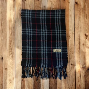Burberry halsduk - Säljer en snygg  halsduk från Burberry i 100% kashmir. Halsduken har ett klassiskt rutmönster i svart, grått och rött samt fransar nedtill. Perfekt för dig som vill ha en stilren och lyxig look inför vintern. Tveka inte att höra av dig med frågor eller funderingar.