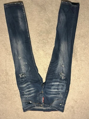 Dsquared2 Ibrahimovic jeans blå - Säljer ett par blå Dsquared2 jeans med tryck 'D2 IBRAHIMOVIC' på låret. Jeansen har slitna detaljer, hål och distressed look samt raka ben. Klassisk femficksmodell med gul kontrastsöm och röd Dsquared2-tag vid gylfen. Snyggt statement-plagg för dig som gillar streetwear.