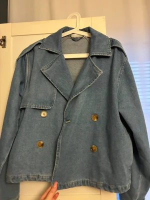 Ljusblå jeansjacka från J.LXX - Säljer en oversized ljusblå jeansjacka från J.LXX i storlek XS. Jackan har dubbelknäppning med stora bruna knappar, bred krage och detaljer vid ärmsluten. Perfekt för dig som gillar en avslappnad och trendig look.