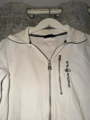 Vit hoodie från Sail Racing - Vit hoodie från Sail Racing med dragkedja framtill och en extra bröstficka med dragkedja. Tröjan har lång ärm, ribbade muddar och diskret logga broderad på bröstet. Tillverkad i mjuk bomull, perfekt för chill dagar.
