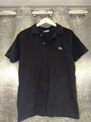 Svart pikétröja från Lacoste - Klassisk svart pikétröja från Lacoste med den ikoniska gröna krokodilloggan på bröstet. Tröjan har korta ärmar, krage och knappar framtill. Tillverkad i mjuk bomull som känns skön mot huden. Perfekt till jeans eller shorts för en clean look.