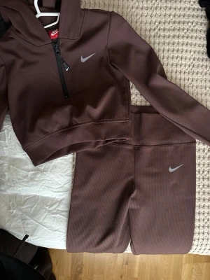 Bruna ribbade Nike tights & hoodie - Säljer ett set från Nike med ribbad struktur i mörkbrun färg. Hoodien har halv dragkedja och Nike-logga på bröstet, medan tightsen har hög midja och matchande logga. Perfekt för träning eller chill. Materialet är stretchigt och mjukt.