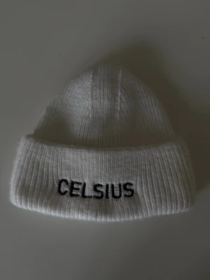 Vit stickad mössa från Celsius - Vit ribbstickad mössa från Celsius med svart broderad logga framtill. Klassisk modell med uppvikt kant som ger en clean och stilren look. Perfekt accessoar för kyliga dagar och enkel att matcha med olika outfits.