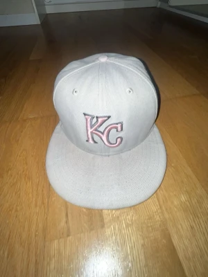 Beige KC keps med broderi - Säljer en beige New Era keps med KC-logga i rosa och silver framtill. Sidan har ett broderat emblem med blommor och KC-sköld. Klassisk rak skärm och broderade ventilationshål. Passar dig som vill ha en clean och unik look.