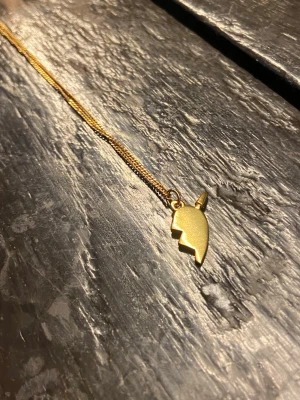 Guldfärgad halsband med blixt-hjärta - Säljer ett guldfärgat halsband med ett coolt blixtformat hjärthänge. Kedjan är tunn och smidig, och hänget har en unik design med en pil som går igenom hjärtat. Perfekt accessoar för dig som vill sticka ut. (Skriv innan köp) 