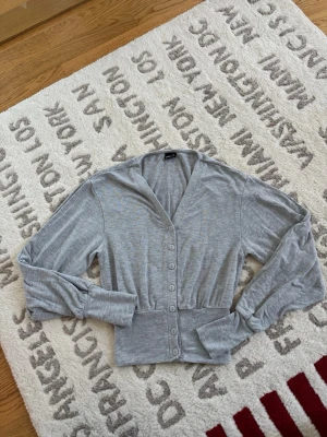 Grå kofta/topp från Gina Tricot XS - Säljer en ljusgrå kofta/topp från Gina Tricot i storlek XS. Koftan har v-ringning, långa ärmar och knappar hela vägen framtill. Tillverkad i mjukt bomullsmaterial och har en ribbad midja som ger en snygg passform.