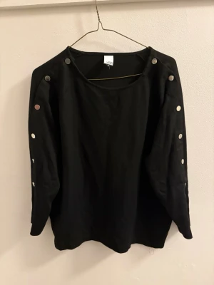 Svart långärmad topp med knappar - En svart långärmad topp med coola silverfärgade knappar längs båda ärmarna. Toppen har en avslappnad passform och är gjord i mjuk bomull, perfekt till jeans eller kjol för en enkel men snygg look.