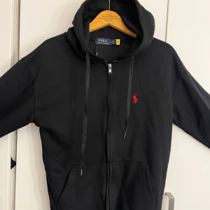 Svart hoodie från Polo Ralph Lauren - Svart hoodie från Polo Ralph Lauren med röd broderad logga på bröstet. Tröjan har huva med snörning, dragkedja framtill och två fickor. Tillverkad i mjuk bomull, perfekt för chill dagar. Klassisk och stilren design som funkar året runt. priset kan diskuteras privat 