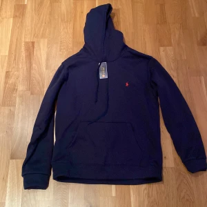 Mörkblå hoodie från Polo Ralph Lauren - Snygg mörkblå hoodie från Polo Ralph Lauren med röd logobrodyr på bröstet. Tröjan har huva, magficka och half zip-dragkedja framtill. Perfekt för chill dagar och enkel att matcha med jeans eller joggers.