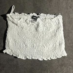 Vit smockad tubtopp från SHEIN - Supersöt vit tubtopp från SHEIN i smockad bomull med volangkant upptill och nedtill. Toppen är axelbandslös och har en bandeauformad siluett som sitter tight mot kroppen. Perfekt för sommarens alla tillfällen, enkel att styla med jeans eller kjol.