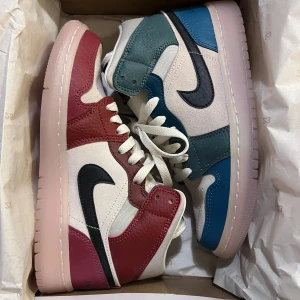 Nike Air Jordan 1 Mid Multicolor - Nike Air Jordan 1 Mid sneakers med unik färgblockning – ena skon i vinrött och vitt, andra i blått och vitt. Båda har svarta swoosh-loggor, vita snören och perforerad tå. Materialet är en mix av skinn och textil, med platt sula och rund tå. Perfekt för dig som vill sticka ut.