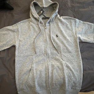 Grå hoodie från Polo Ralph Lauren - Snygg grå hoodie från Polo Ralph Lauren med dragkedja och huva. Tröjan har snörning vid huvan, ribbade muddar och fickor framtill. Klassisk Polo-logga broderad på bröstet. Helt ny med taggar Endast provad Lite mindre i storleken
